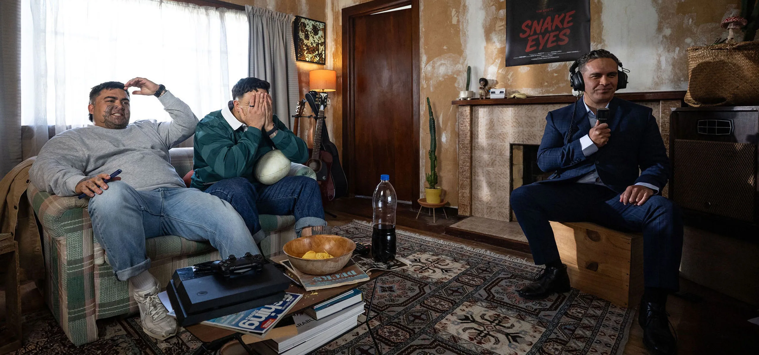 A commentator in a suit calls the action like a sports broadcast from a living room, while two men watch from the couch, one with his head in his hands.